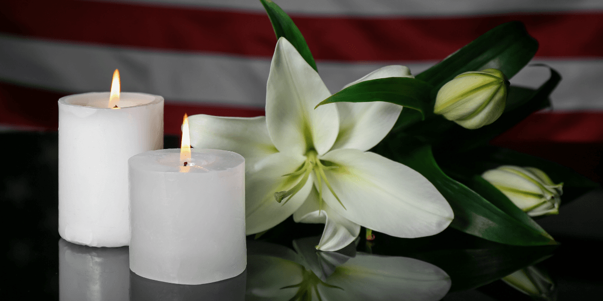 Two lit candles beside a white lily arrangement on a reflective surface