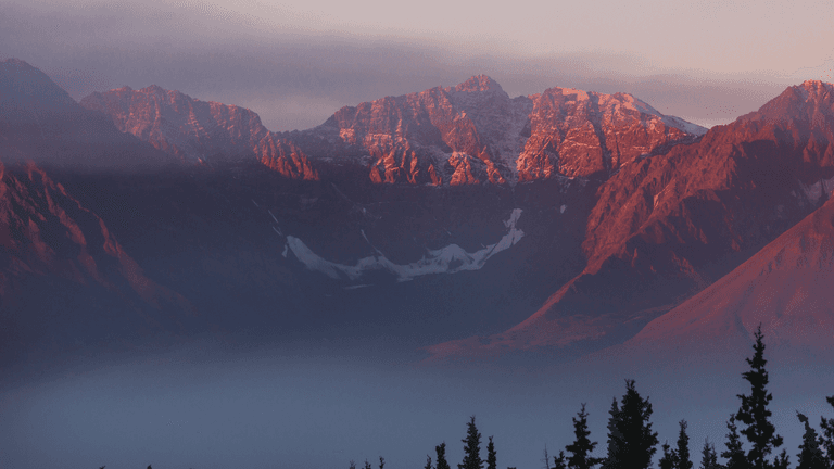 Sunrise view of a mountain landscape with rugged peaks, low clouds, and evergreen trees in the foreground.