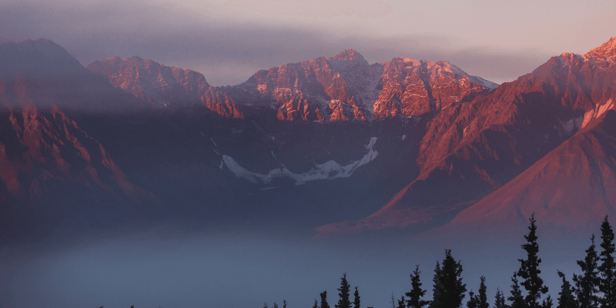 Sunrise view of a mountain landscape with rugged peaks, low clouds, and evergreen trees in the foreground.