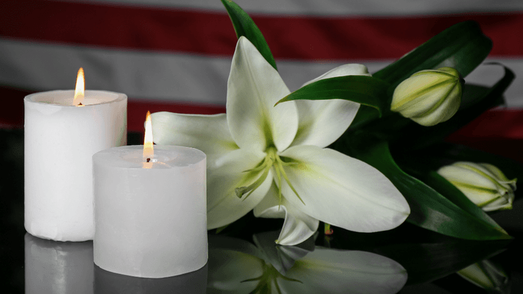 Two lit candles beside a white lily arrangement on a reflective surface