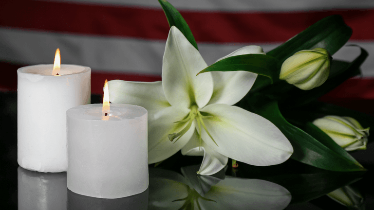 Two lit candles beside a white lily arrangement on a reflective surface
