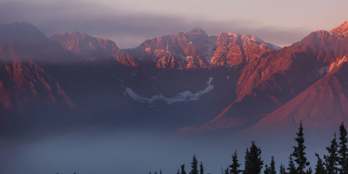 Sunrise view of a mountain landscape with rugged peaks, low clouds, and evergreen trees in the foreground.